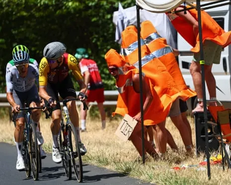 Three cyclists ride past spectators in orange clothing waving flags along a sunny roadside