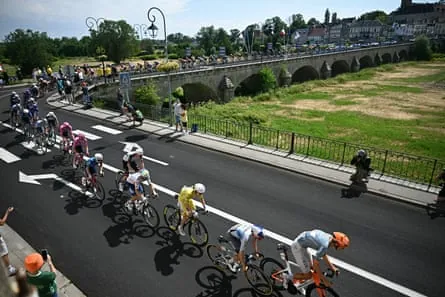 Tadej Pogacar and the peloton cross a bridge on the dried-up riverbed of La Vieille Loire river