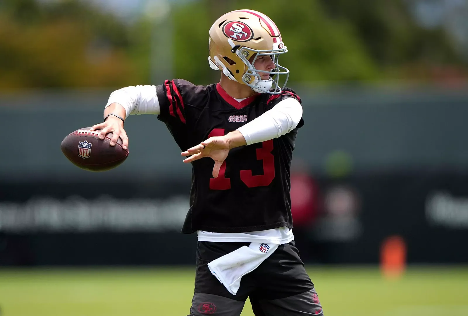 A football player wearing a San Francisco 49ers helmet and black jersey with number 13 prepares to throw a pass on a grassy field.